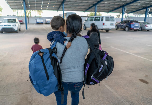 A mother holding a toddler and two children carrying bags walking in a parking lot. 