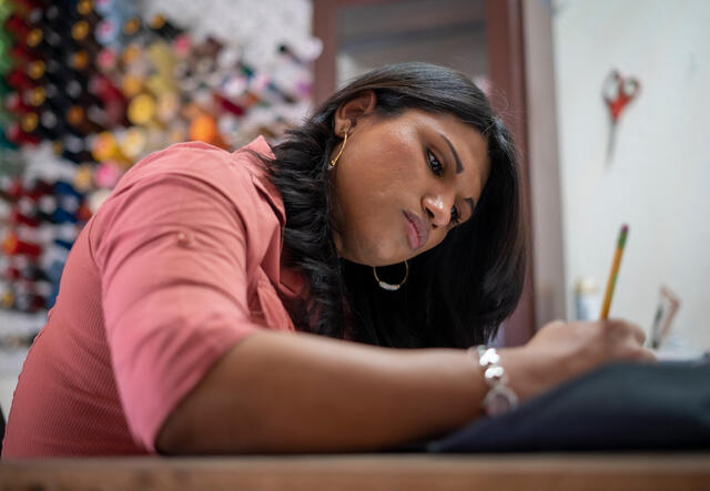 Lincy Sopall, who was granted asylum in the U.S., sits at a desk holding a pencil and looking at a sheet of paper. Her supplies for her fashion design studio are behind her.