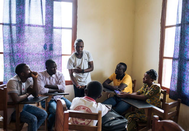 Benjamin, standing in a classroom in Congo, speaks to other teenagers seated at desks around him about Ebola. 