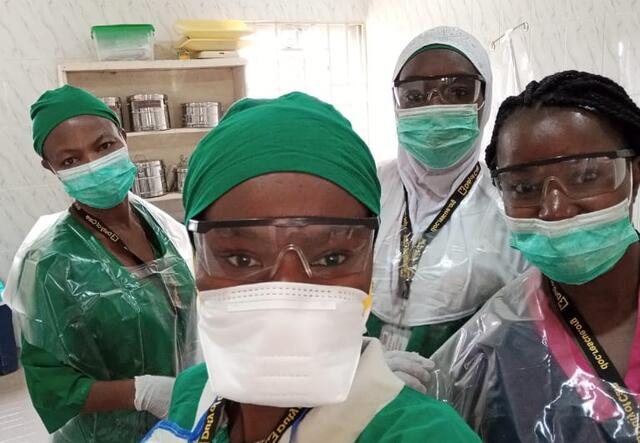 Wearing green scrubs, masks and face shields, midwives in an IRC reproductive health clinic take a selfie