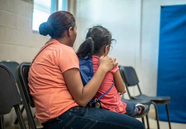 In an IRC shelter, a young mother sits with her back to the camera and her arm around her 4-year-old daughter. 