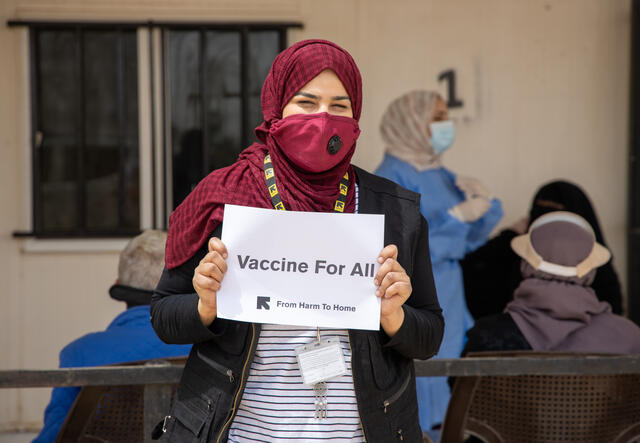 An IRC senior health officer wearing a face mask against COVID-19 stands holding a "Vaccines for All" sign at the IRC health clinic where she works in Zaatari refugee camp in Jordan