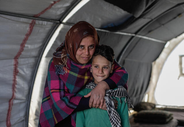 Sundus hugs her son Omar while standing behind him in a tent. Both are smiling and looking at the camera. 