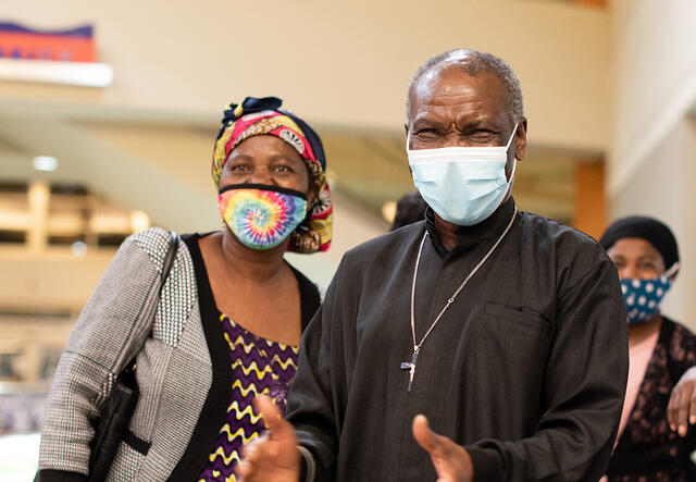 Patrice and Wanyema, both wearing masks, both watch their daughter arrive at the Boise airport. Patrice is about to clap his hands. 