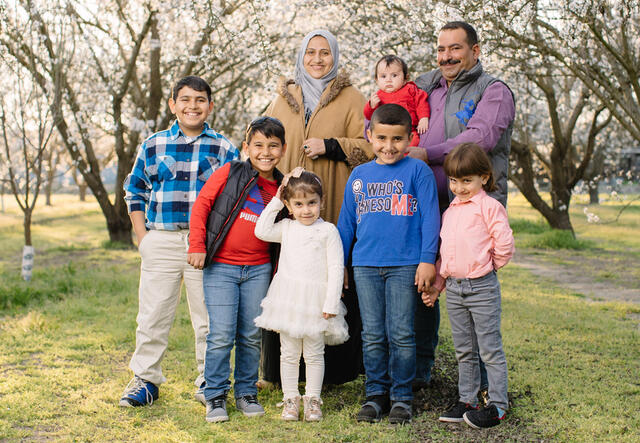 A husband and wife from Syria and their six children are standing and smiling in the middle of an almond orchard when the almond trees are blooming.