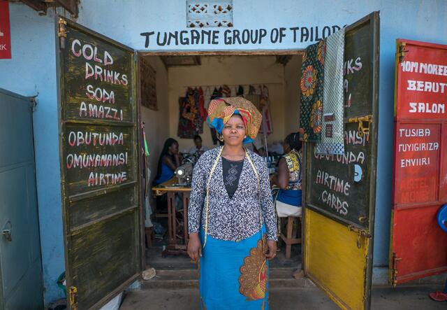 Domitila Kaliya, a Congolese refugee living in Kampala, Uganda, stands with a proud smile outside her shared tailoring workspace.