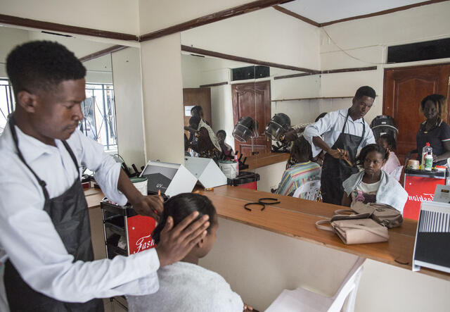 A man styles a seated woman's hair in front of a large mirror during a hairdressing training class.