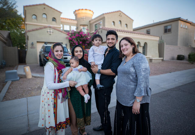 In front of a house, Muska Haseeb holds a baby and poses with her family, one man, two other woman and two babies. 