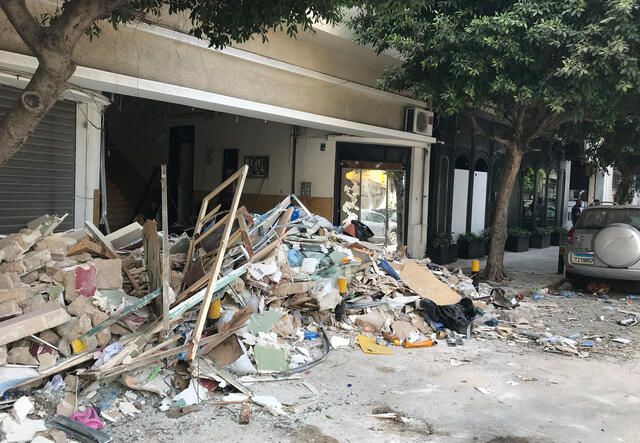 A building on a tree-lined city street. One of the buildings has no front wall and there is a pile of rubble in front of it. 