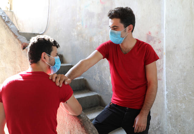 21-year-old Barakat, a Syrian refugee in Lebanon, stands on a stair wearing a mask with his hand on the shoulder of his brother Mohammed. They are wearing matching shirts. 