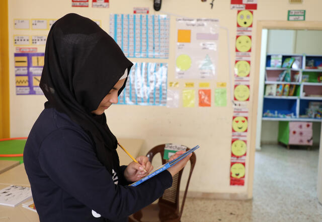 A young girl stands and writes in a book she is holding. She is in a brightly-colored room full of educational materials. 