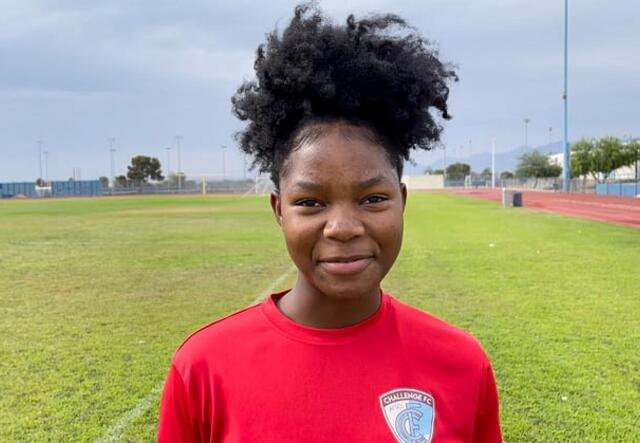 Wearing a red shirt and smiling, Kezia looks at the camera while on a soccer field 