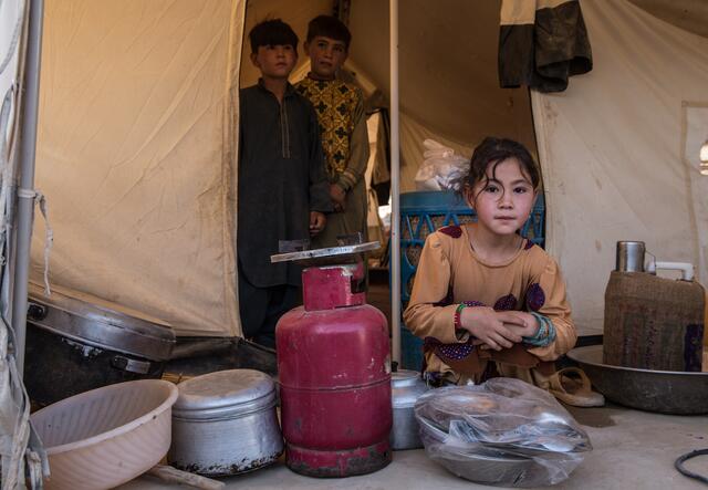 A displaced Afghan girl crouches outside her tent in Afghanistan with two of her brothers standing behind her in the doorway.