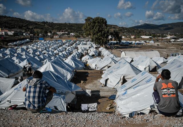 Two refugees sit looking out over a sea of white tents in a refugee camp on Lesbos, Greece.