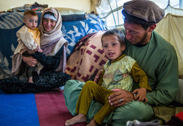 A mother and father in Afghanistan sit on the ground among blankets and pillows, each with a child on their lap. The boy in the front is smiling and his father is looking at him. 