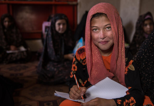 A young girl sits with a notebook on her lap ready to write while looking up at a teacher. There are other young girls sitting behind her. 