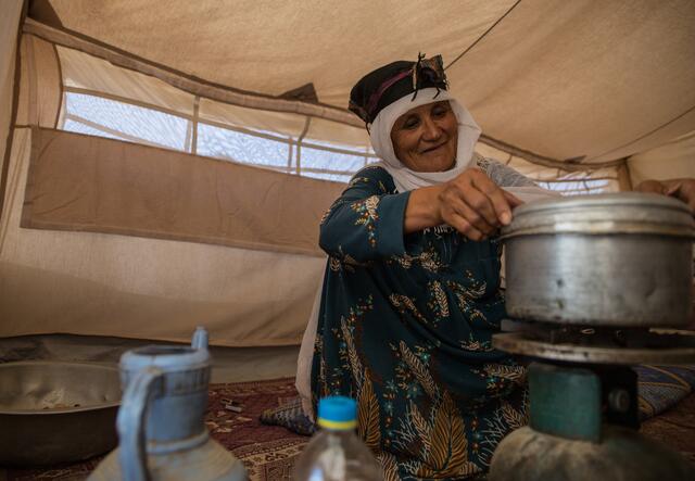 An elderly Afghan woman crouches on the floor of a tent in a camp in Badghis, Afghanistan cooking a meal in an aluminum pot on a burner.