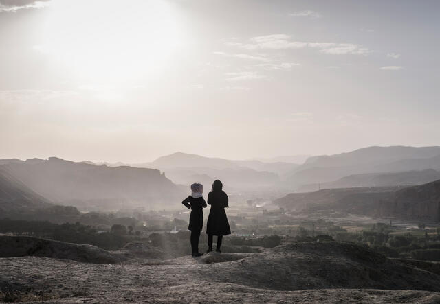 A landscape photo with a mountainous landscape and the silhouettes of two girls who are looking at the landscape with their backs to the camera.  