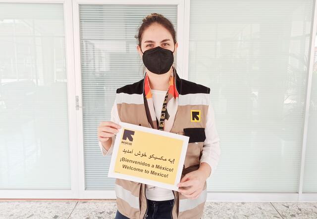 Fatima, a home visiting officer for the IRC in Mexico, holds a sign that says "welcome to Mexico" in English, Spanish and Dari. She is wearing a mask and an IRC vest. 
