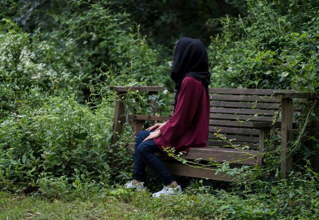 Afghan refugee Fatima, 19, sits facing sideways on a wooden garden swing in a lush garden in Richmond, VA
