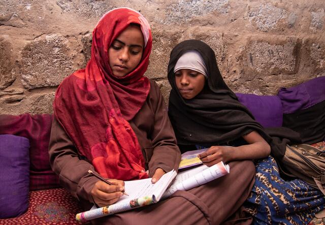 In a camp for displaced families in Yemen, Na'aem, 11 and Aisha, 10, sit on the ground going over a school workbook together and writing. The girls are best friends.