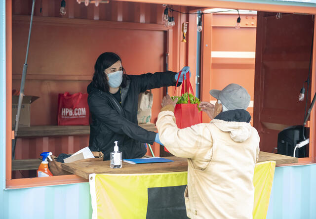 A woman wearing a mask hands a tote bag of food to a refugee.