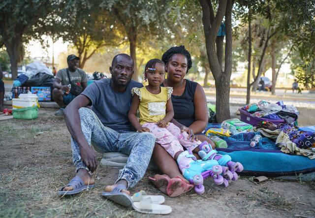 In a makeshift encampment in Mexico, a Haitian family--a mom, dad and young daughter--look straight at the camera while sitting on the ground next to their suitcases and blankets. 