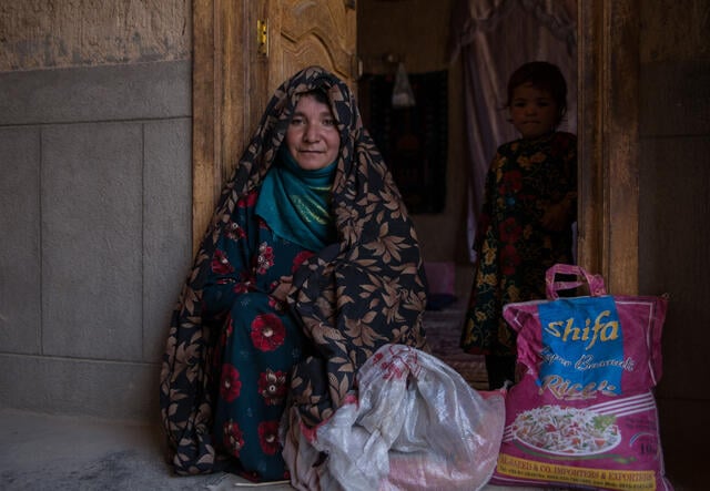 An Afghan mother sits next to large sack of rice with her toddler standing in the doorway behind her.