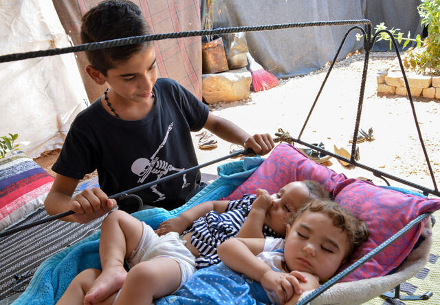 A Syrian boy rocks two young children in a cradle.