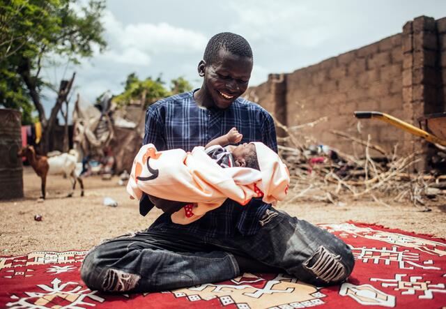 A Nigerian man smiles and holds his baby while sitting on a rug.