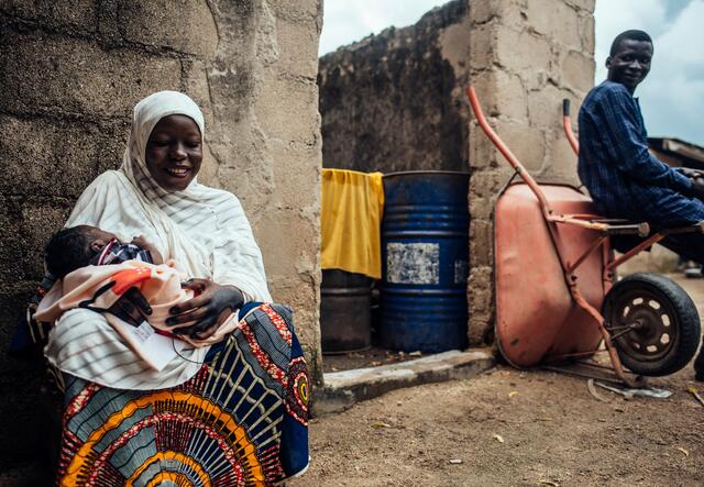 A Nigerian woman smiles while holding her baby and being looked at by husband who sits on a wheelbarrow.