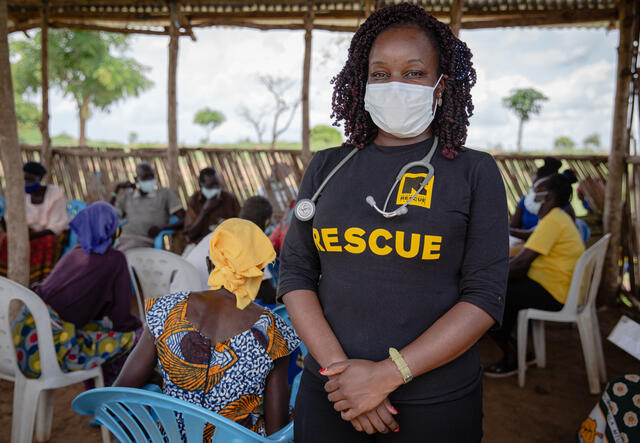 Dr. Ruth Grace Babirye stands wearing a protective face mask as elderly people  sit in an open waiting room in Bidi Bidi refugee settlement in Uganda to get vaccinated against COVID-19. 