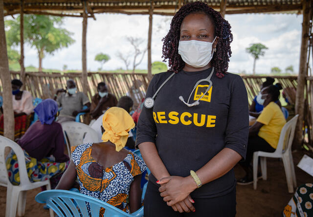 Dr. Ruth Grace Babirye stands wearing a protective face mask as elderly people sit in an open waiting room in Bidi Bidi refugee settlement in Uganda to get vaccinated against COVID-19.