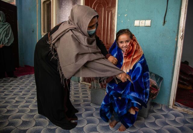 Noor bends over to adjust a blue blanket her daughter has draped over her shoulders. Her daughter is smiling. 