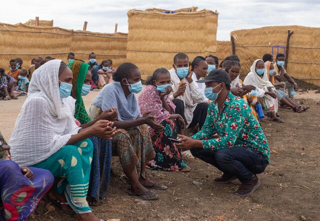 People in the Tuneiba camp in Sudan sit on benches and a man kneels in front of them. 