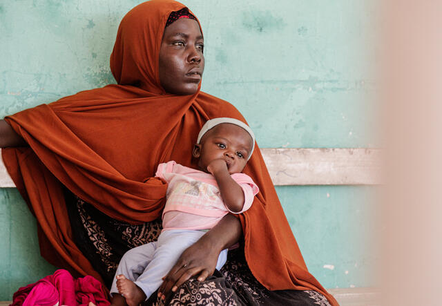 A woman sits against a wall with her baby on her lap