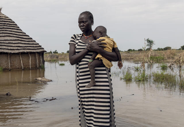 With water above her ankles, a mother holds her child and stands in front of her flooded home. 