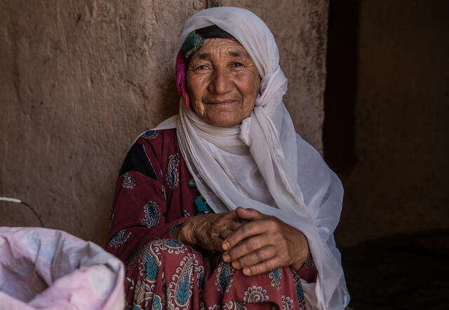 An older woman sits on the ground next to a bag of grain. 