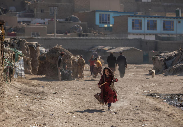 A girl runs through a camp in a desert landscape. There are homes, other children playing and adults walking behind her. 