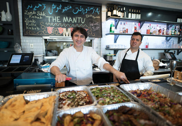 Smiling, a chef and his employee stand behind large trays of food with a chalkboard behind them that says "The Mantu." 