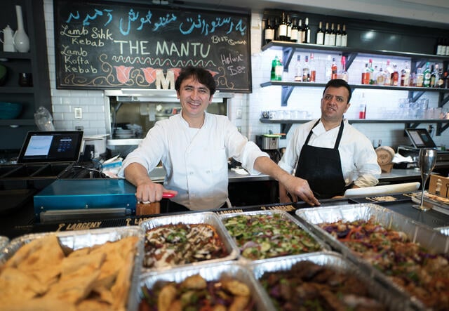 Smiling, a chef and his employee stand behind large trays of food with a chalkboard behind them that says "The Mantu."