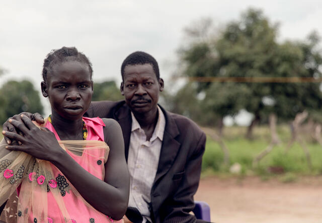 A man an a woman sit next to each other outside. He has a hand on her shoulder and they are looking at the camera. 