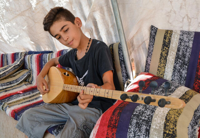 In a tent, a 13-year-old boy sits and plays the buzuq. 
