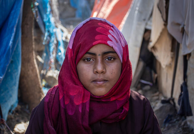 A portrait of Na'aem, a young girl who is looking straight at the camera and wearing a pink scarf. There are tents and makeshift shelters behind her. 