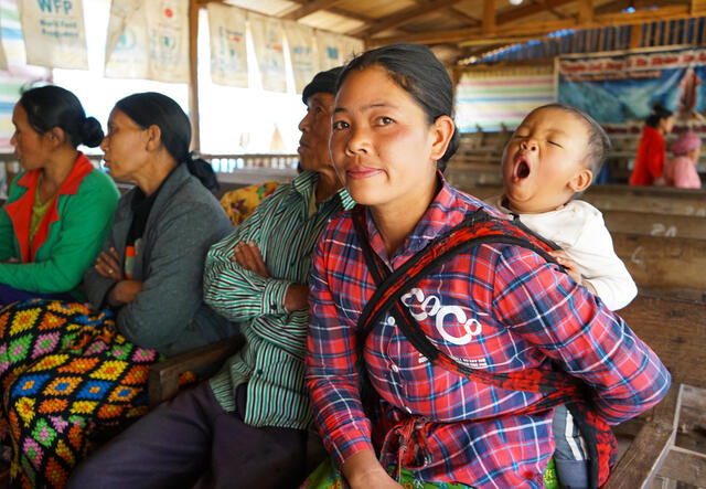 A young Shan mother sits in a bamboo structure alongside other women, her baby yawning while nestled in a sling on her back.