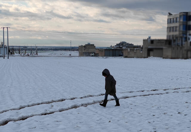 A person wearing a winter coat and boots treks through the snow past a decrpit building in Syria.