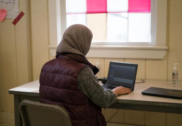 A young woman, with her back to the camera, sits at a desk and types on her laptop.