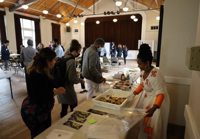 A woman standing behind a table serves food to a line of three people. In the background is an auditorium with more people mingling with each other, out of focus.