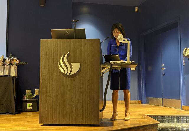 A woman in a blue dress reading from behind a podium decorated with the GSU logo.
