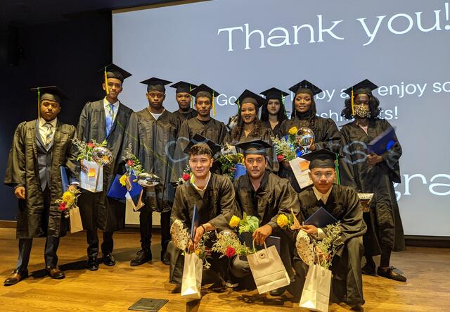 A group of 12 graduates on stage and in their robes posed for a photo. There is a projection cast over them that reads "Thank you! Go to the lobby and enjoy some refreshments. Congrats grads!"